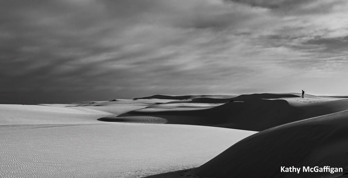 A black and white shows a moody scene of large sand dunes with a person standing in the distance.
